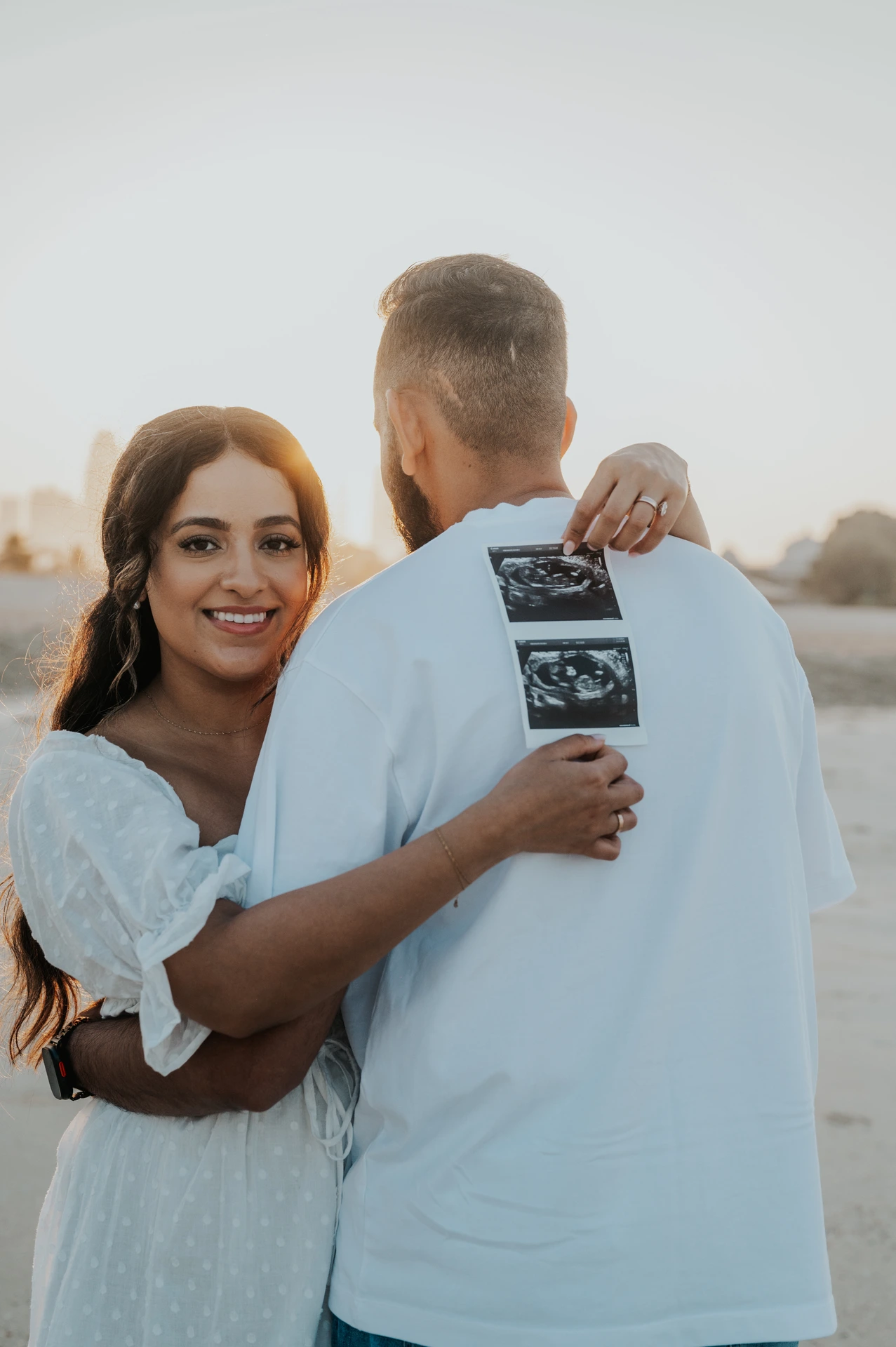 A mother-to-be smiles while holding ultrasound prints against her partner’s back on Dubai beach during a maternity photoshoot.