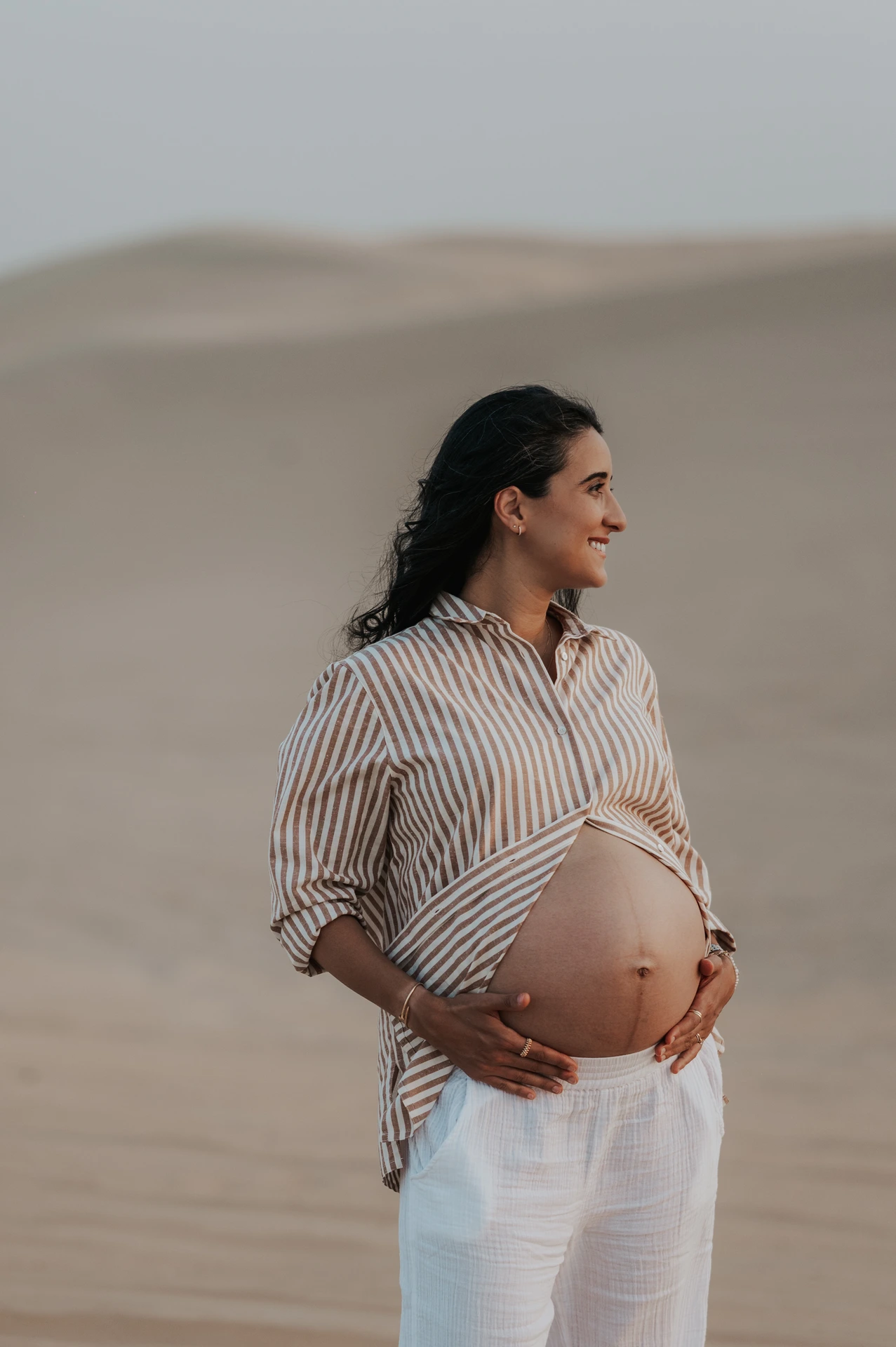Maternity photoshoot Dubai - striped shirt baby bump A pregnant woman in a striped shirt reveals her baby bump on Dubai beach during a maternity photoshoot with warm natural light.