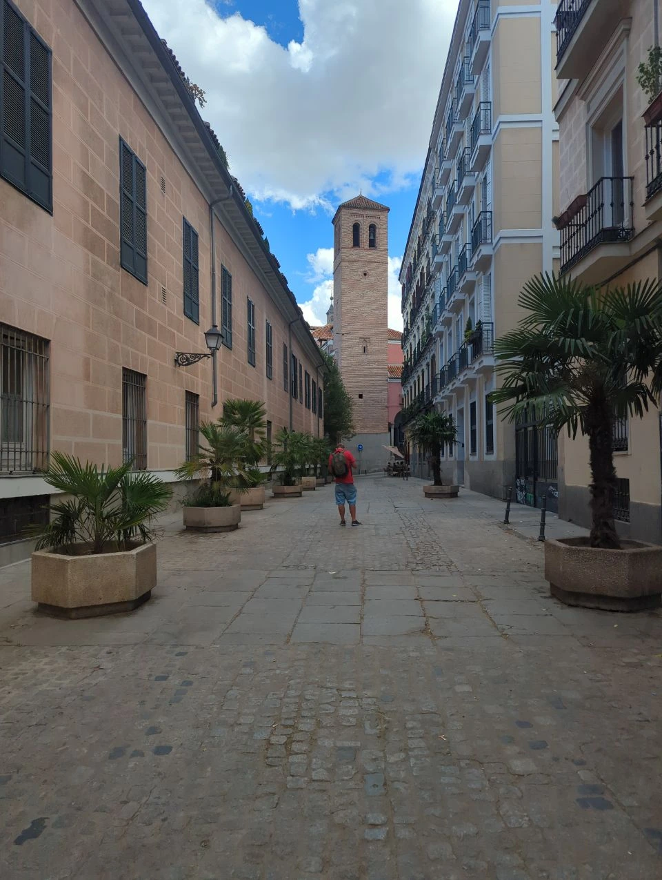 Madrid Midday Photoshoot - Historic Lane A pedestrian walks along a quiet cobbled lane in La Latina, framed by warm facades, palms and a brick tower in Madrid.
