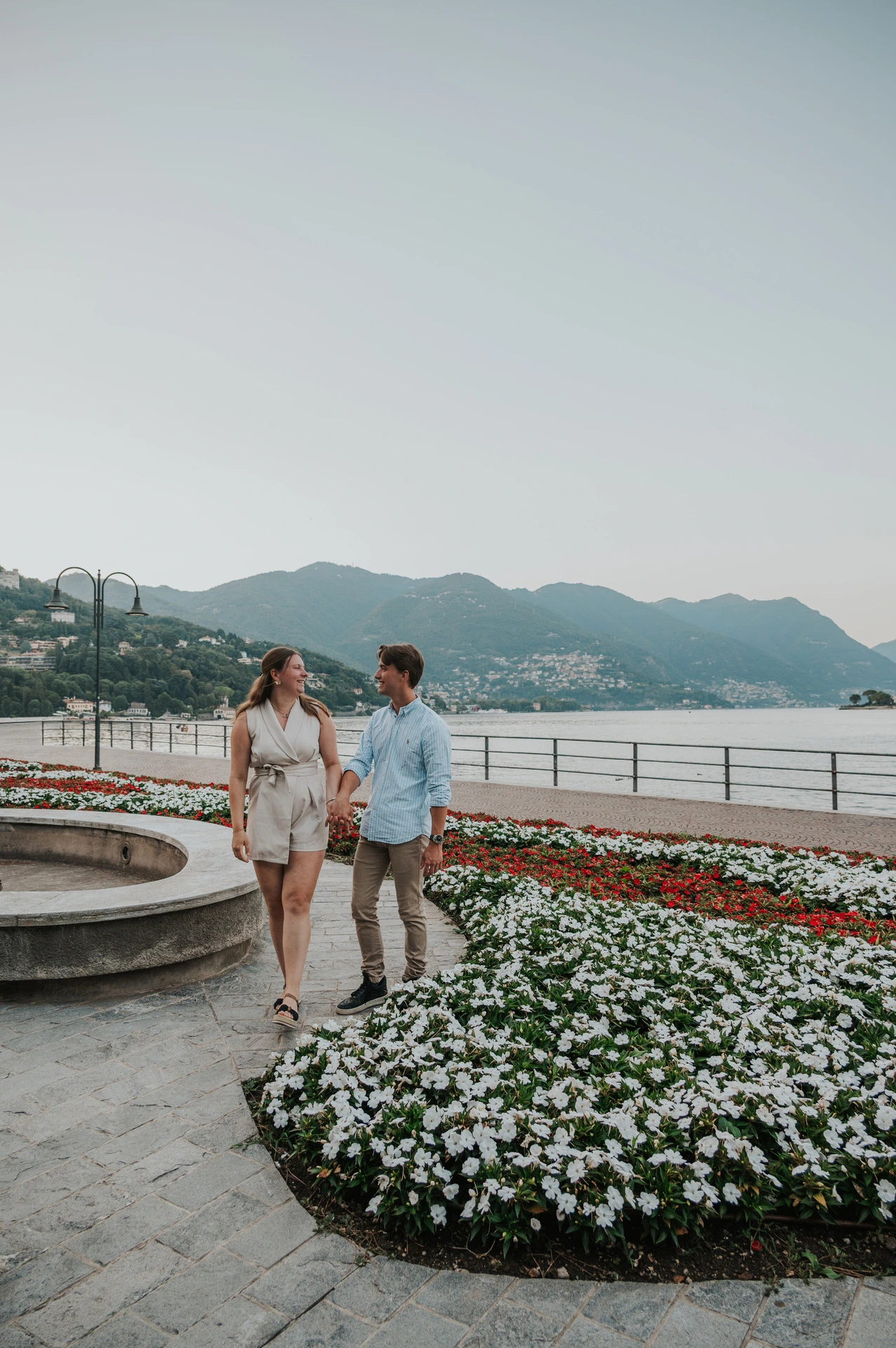 Couple walking beside a fountain and red and white flowers during their Lake Como couple photoshoot on Como’s promenade.