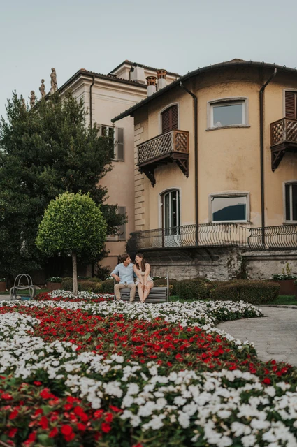 Lake Como couple photoshoot among Como’s flower beds Couple walking beside red and white flower beds with a yellow building behind them during their Lake Como couple photoshoot.