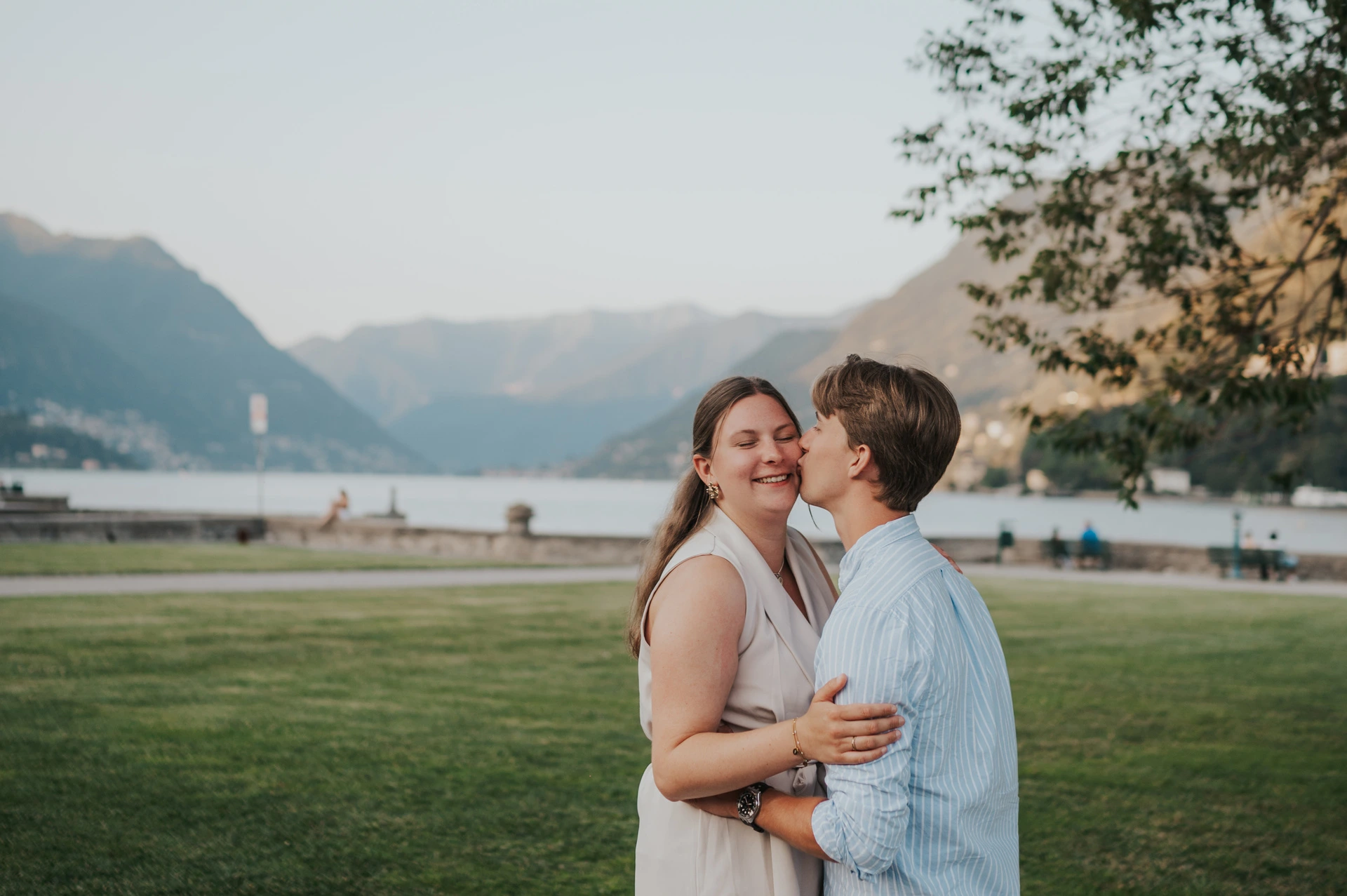 Man kissing his partner’s cheek on the lawn beside Lake Como during their couple photoshoot in the gardens of Villa Olmo.