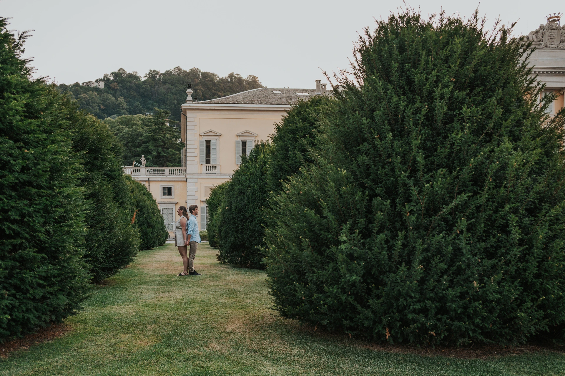 Wide view along a path between clipped hedges towards the couple during their Lake Como couple photoshoot at Villa Olmo.