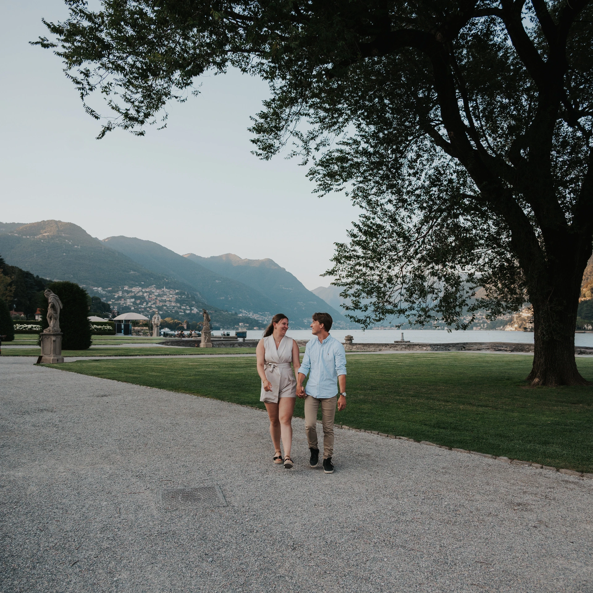 Couple walking along a broad garden path beneath a leafy tree during their Lake Como couple photoshoot at Villa Olmo.