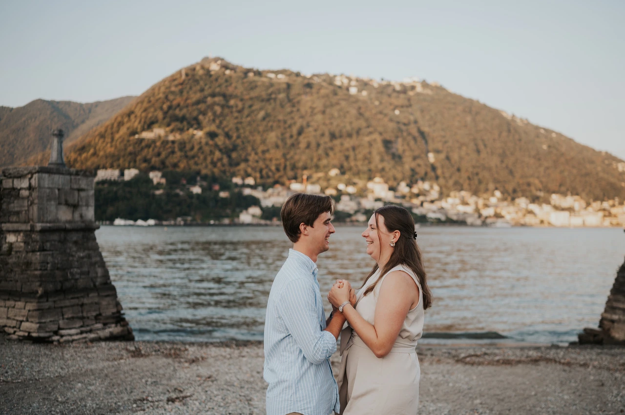 Lake Como couple photoshoot by the gardens’ lakeside wall Couple embracing beside a low stone wall during their Lake Como couple photoshoot, with hillside houses across the lake.
