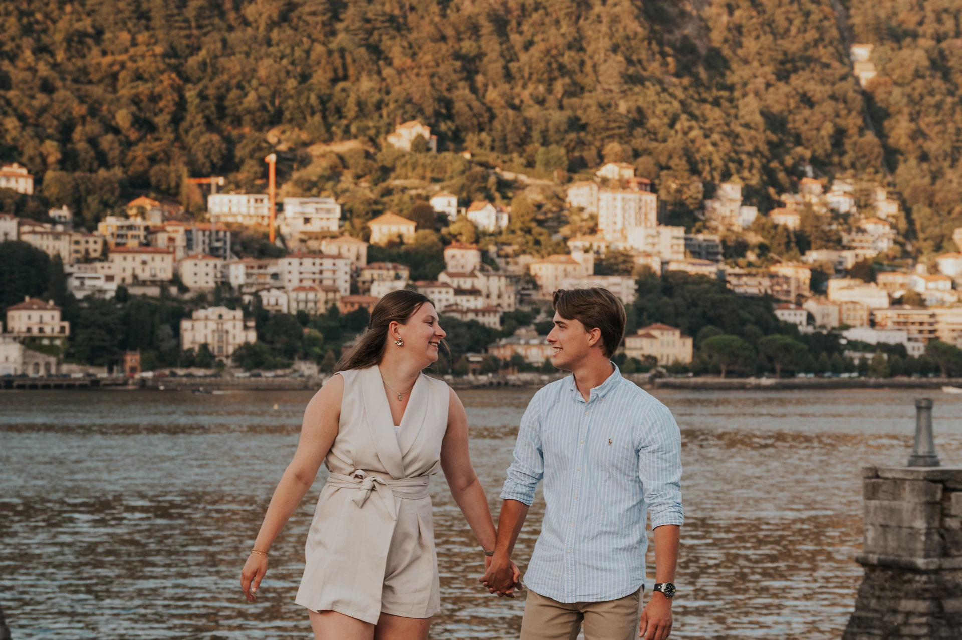 Couple walking hand in hand beside a stone wall during their Lake Como couple photoshoot, with water and hillside houses behind them.