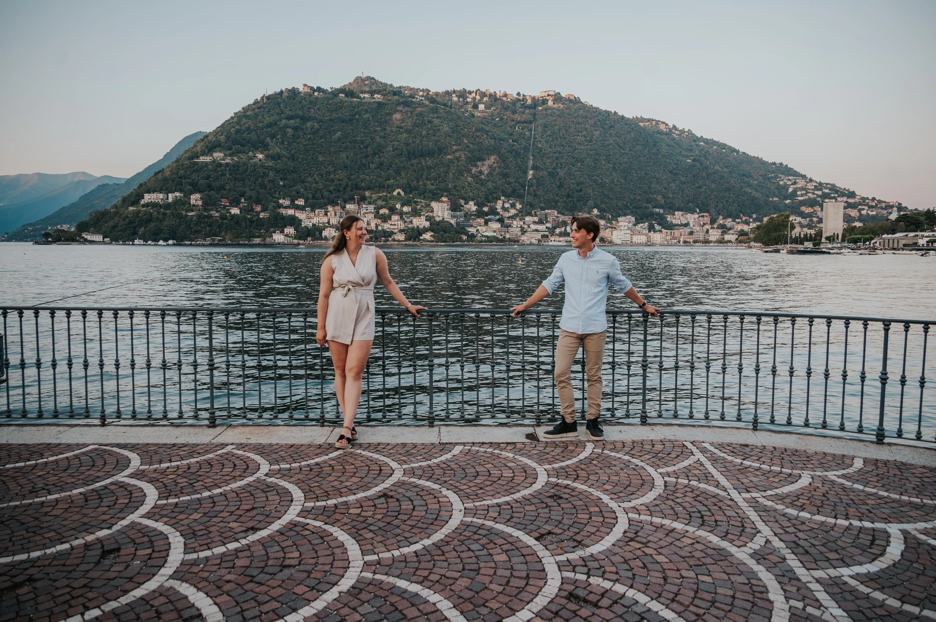 Couple standing apart by a curved waterfront railing during their Lake Como couple photoshoot, with patterned paving in the foreground.