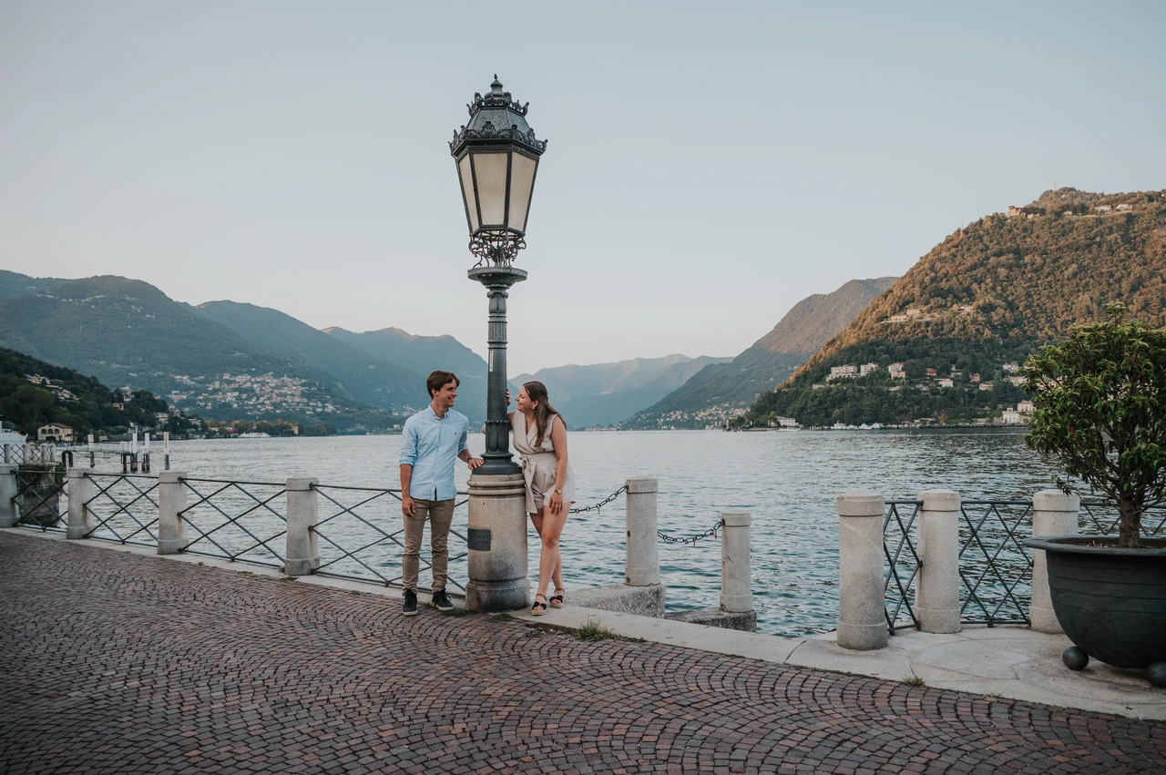 Lake Como couple photoshoot beside a waterfront lamp Wide view of the couple beside a lamp on Como’s waterfront during their Lake Como couple photoshoot, with mountains across the lake.