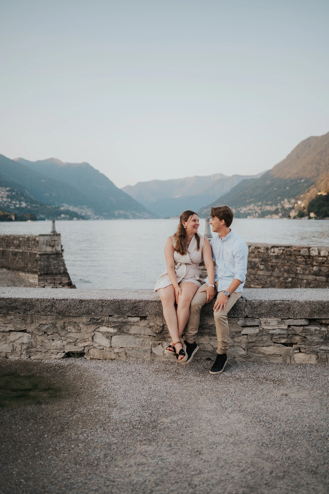 Couple sitting together on a stone wall on Como’s promenade during their Lake Como couple photoshoot, with mountains across the water.