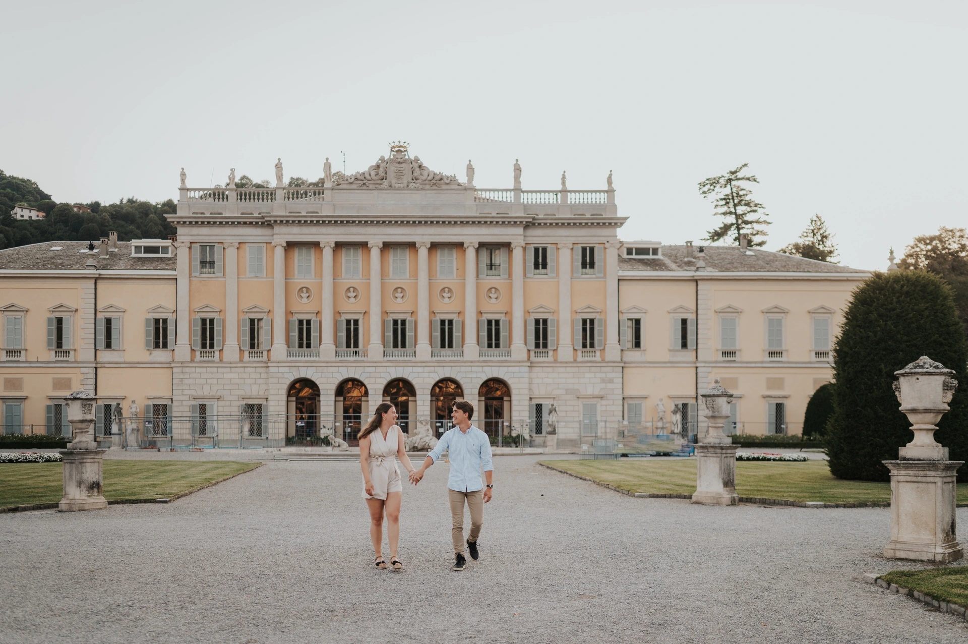Lake Como couple photoshoot with Villa Olmo’s full facade Wide view of the couple walking towards the camera in front of Villa Olmo during their Lake Como couple photoshoot.