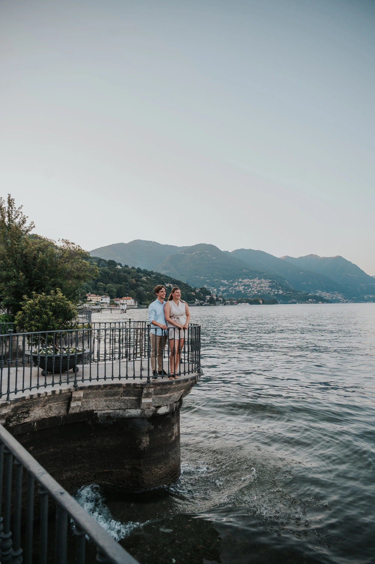 Wide view of the couple on a railed lookout over the water during their Lake Como couple photoshoot in Como.