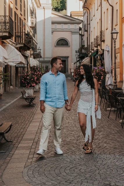 Lake Como engagement photoshoot through the streets of Menaggio The couple walk together along a cobbled Menaggio street between warm-coloured buildings during their Lake Como engagement photoshoot.