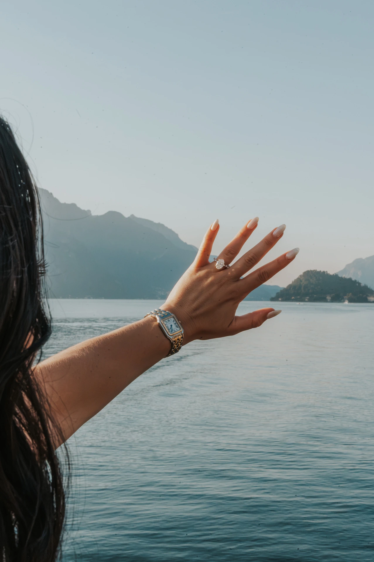 The woman raises her left hand to show her engagement ring against the water and mountains during their Lake Como photoshoot in Menaggio.