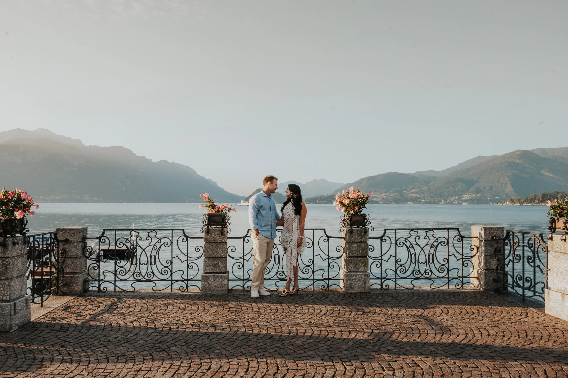 A wide photograph of the couple on a cobbled terrace framed by flowers, lake and mountains during their Lake Como photoshoot in Menaggio.