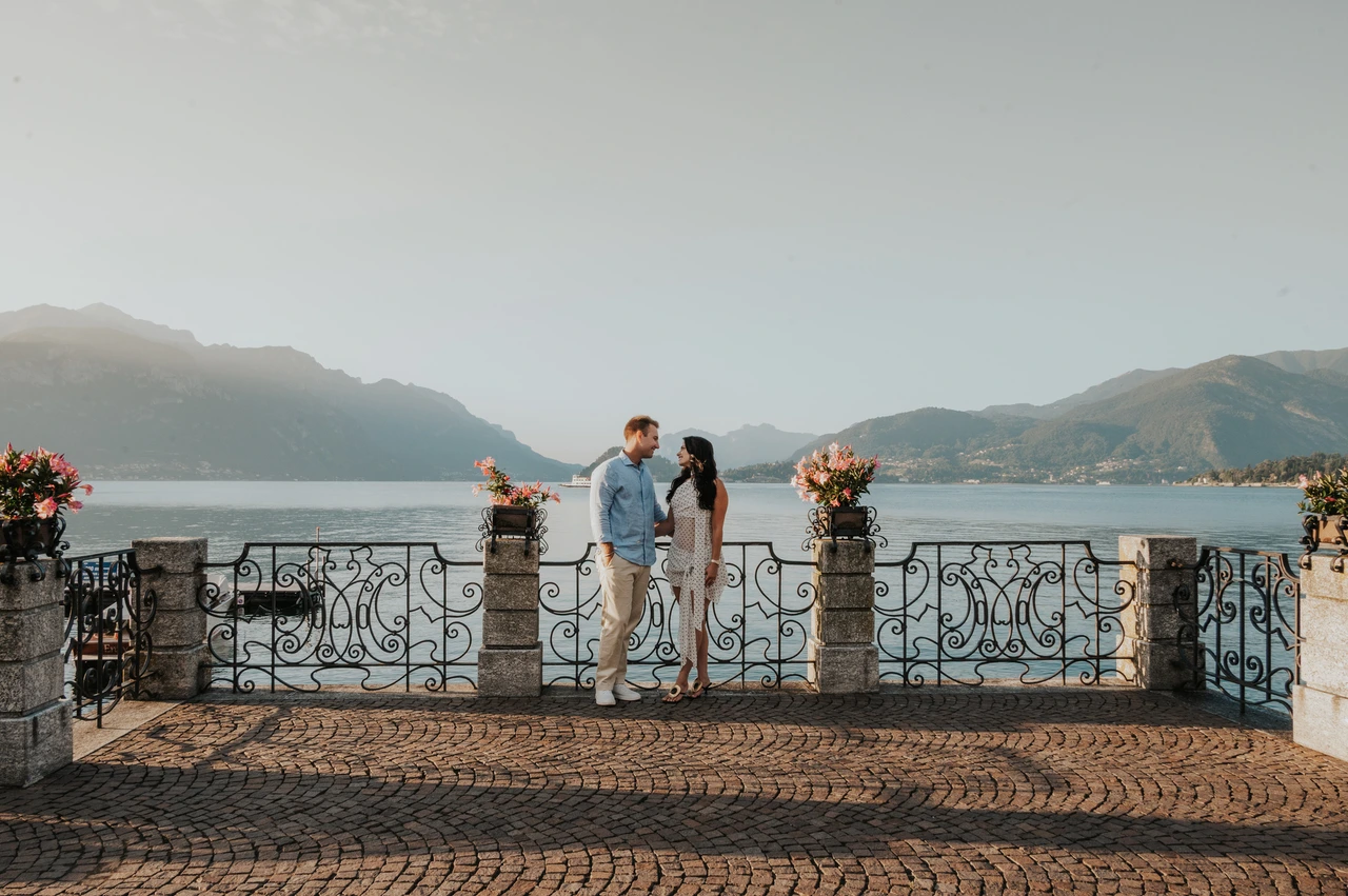 Wide Lake Como photoshoot on Menaggio’s flower-lined terrace A wide photograph of the couple on a cobbled terrace framed by flowers, lake and mountains during their Lake Como photoshoot in Menaggio.