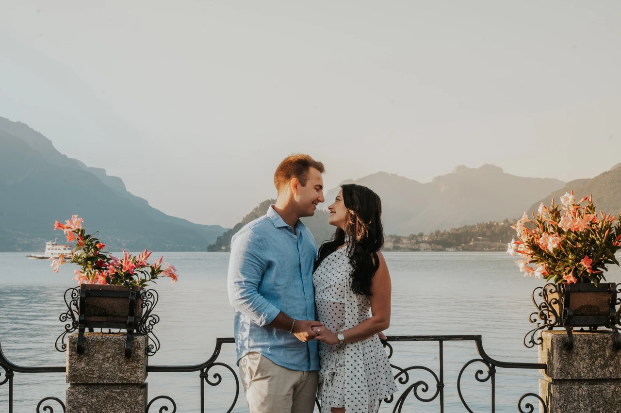 Lake Como engagement photoshoot framed by pink flowers in Menaggio The couple smile at each other between pink flowers, with the lake and mountains behind them during their Lake Como engagement photoshoot.