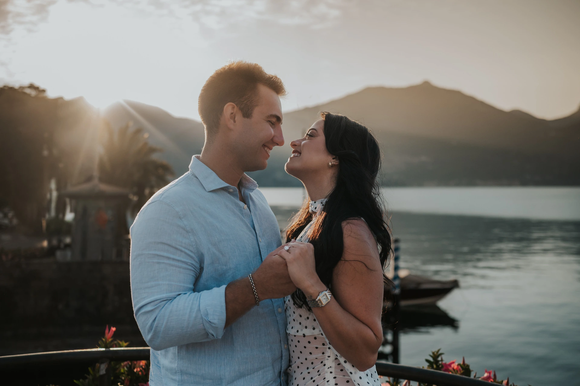The newly engaged couple hold hands beside the water, with light over the mountains behind them during their Lake Como photoshoot in Menaggio.