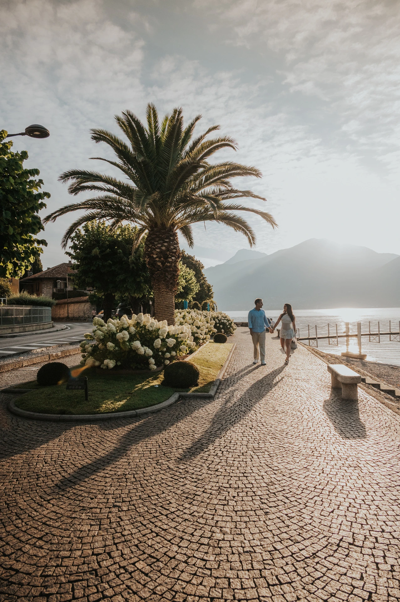 The couple walk beside the water under a tall palm, with cobbles and a stone bench in view during their Lake Como photoshoot in Menaggio.