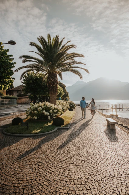 Lake Como photoshoot on the Menaggio promenade beneath a palm The couple walk beside the water under a tall palm, with cobbles and a stone bench in view during their Lake Como photoshoot in Menaggio.