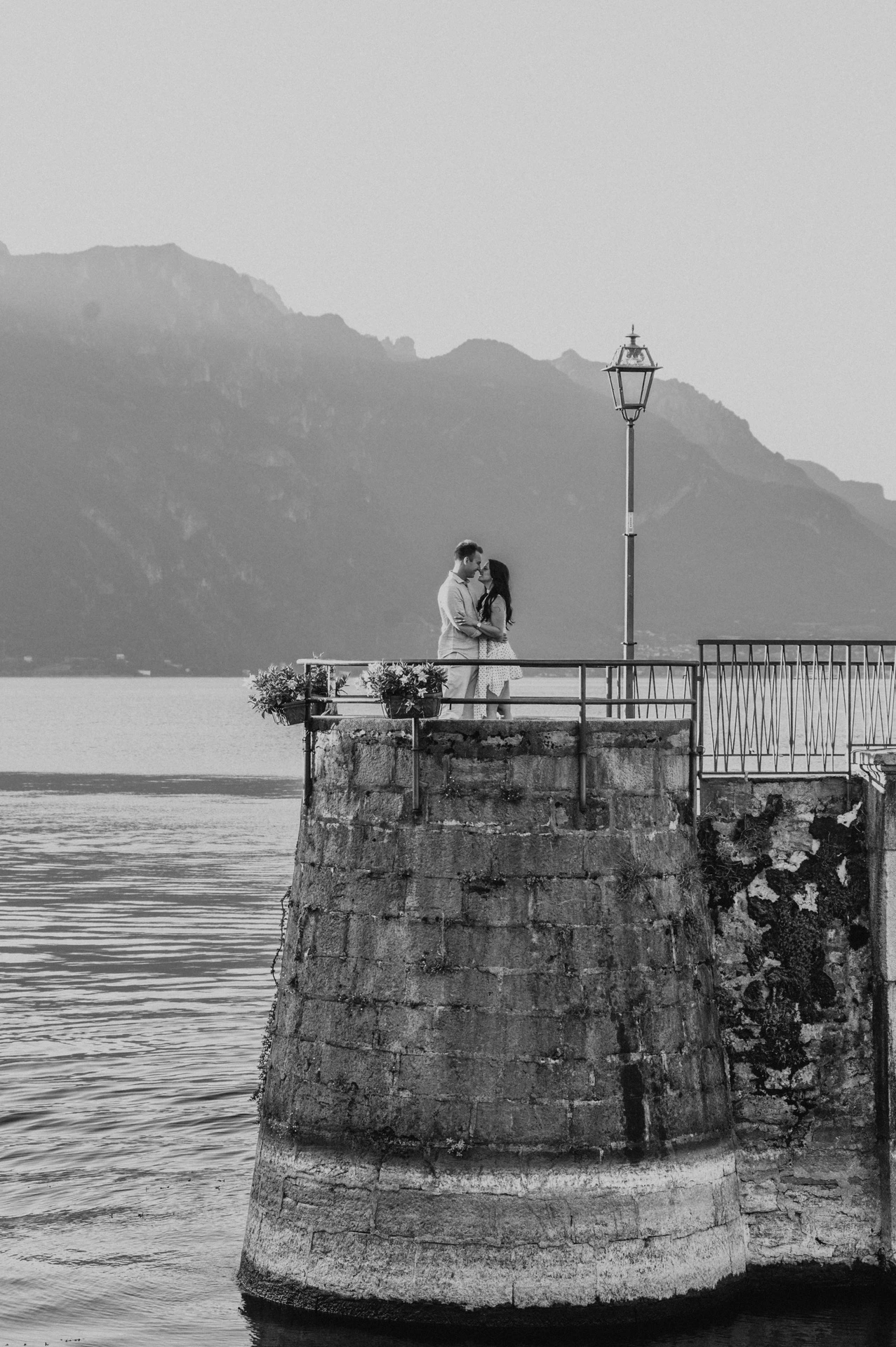 Black-and-white photograph of the couple on Menaggio’s rounded stone quay, with mountain slopes across the lake during their Lake Como photoshoot.