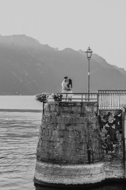 Black-and-white Lake Como photoshoot on the rounded Menaggio quay Black-and-white photograph of the couple on Menaggio’s rounded stone quay, with mountain slopes across the lake during their Lake Como photoshoot.
