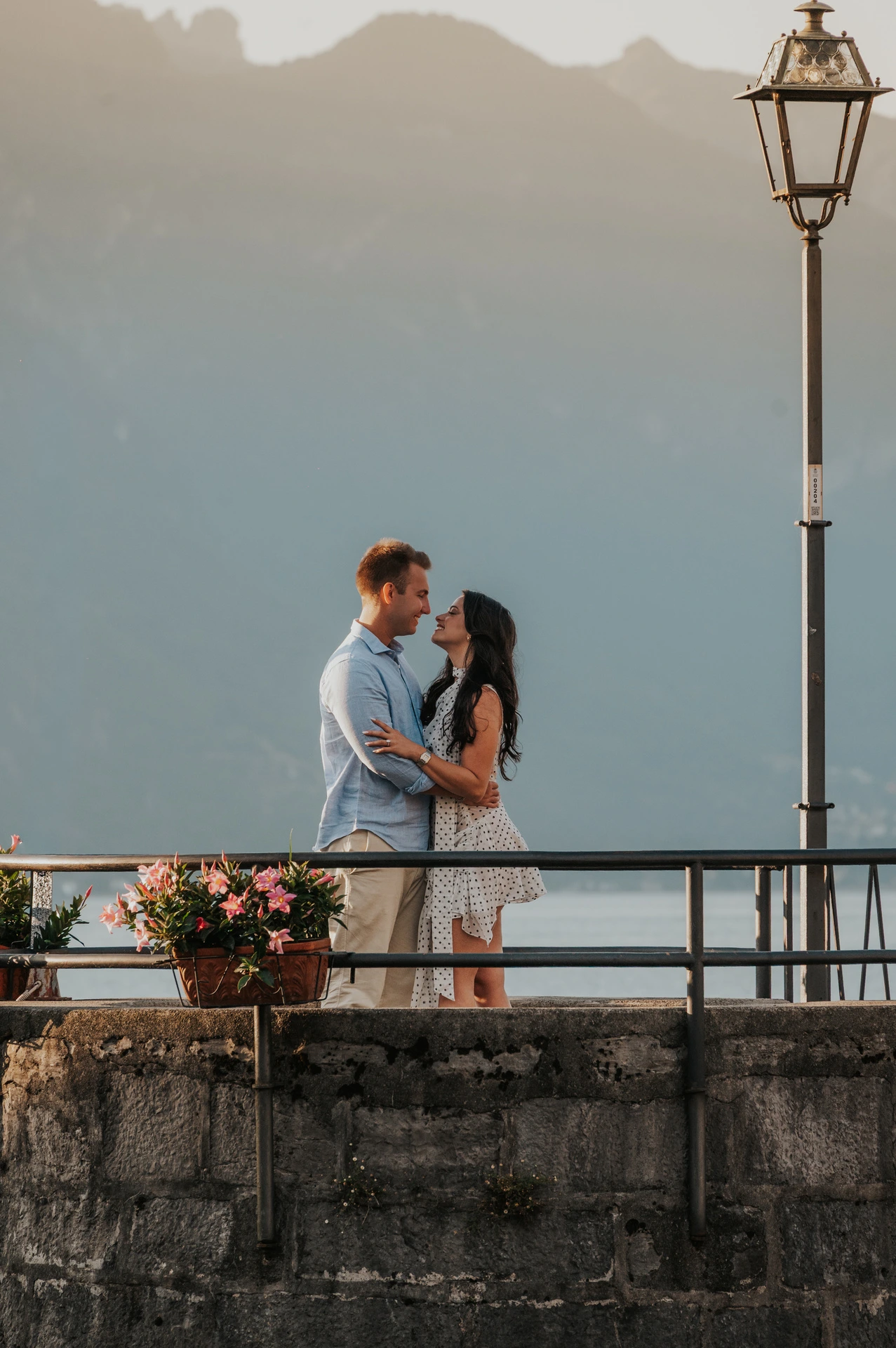 The couple stand close beside a tall lamp and flowers on Menaggio’s stone quay during their Lake Como photoshoot.