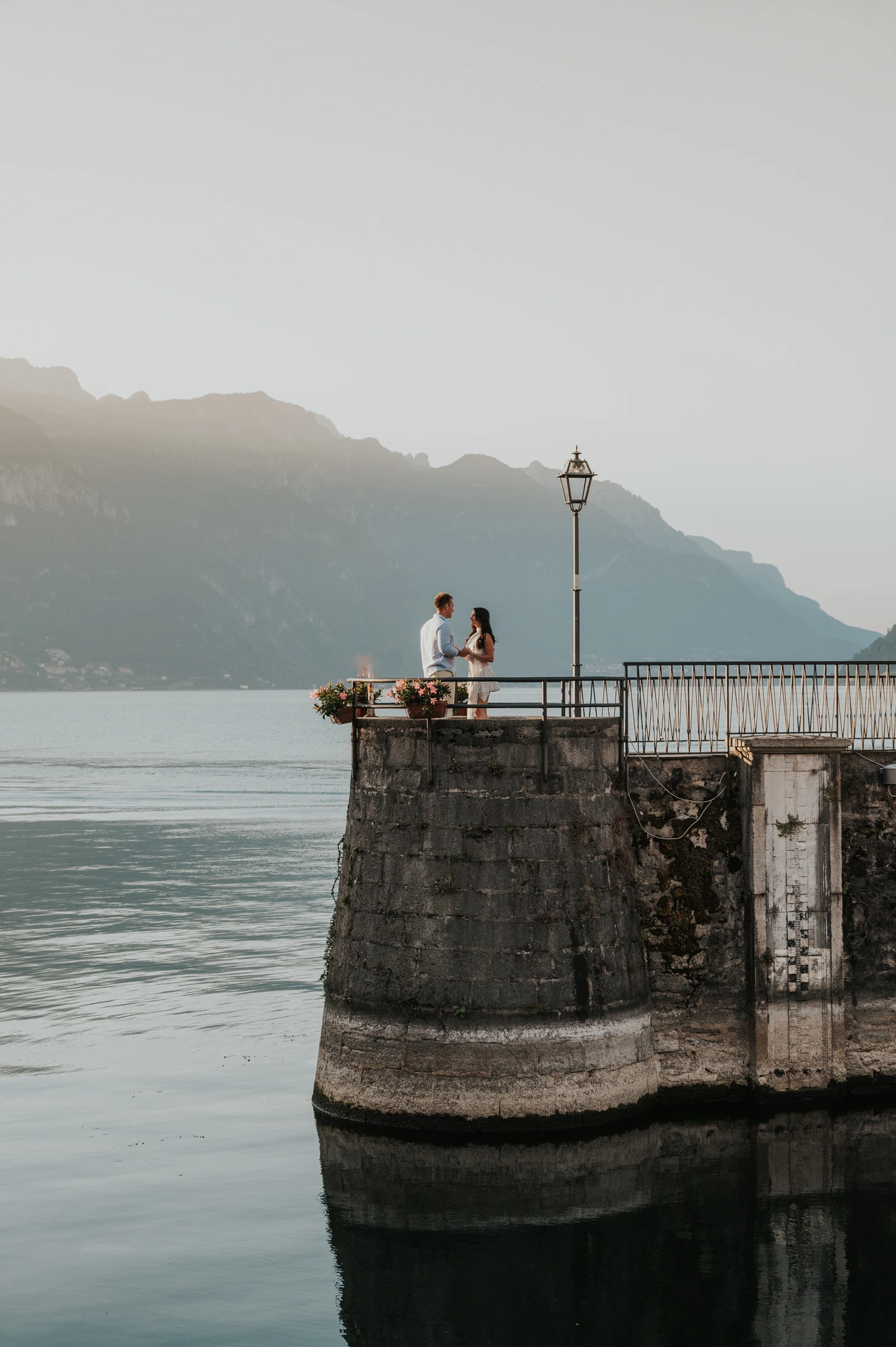A wide view of the couple on a rounded stone quay above the water during their Lake Como engagement photoshoot in Menaggio.