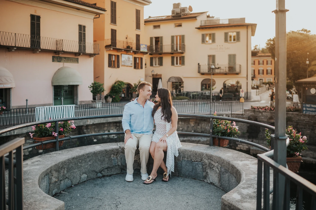 Lake Como engagement photoshoot beside Menaggio’s colourful buildings The couple sit together on curved stone seating beside the colourful buildings of Menaggio during their Lake Como engagement photoshoot.