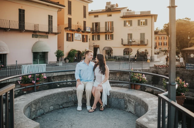 The couple sit together on curved stone seating beside the colourful buildings of Menaggio during their Lake Como engagement photoshoot.