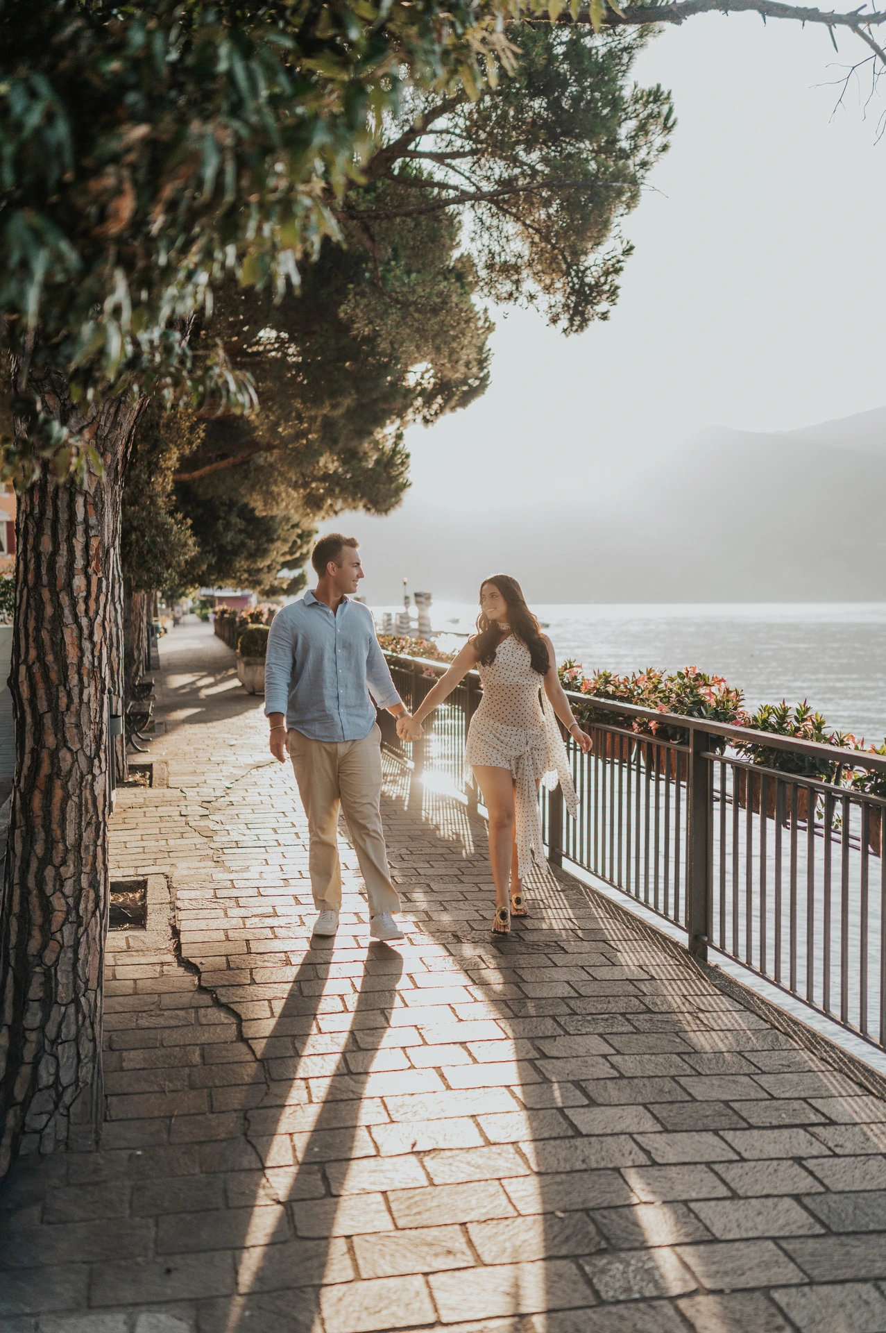 The couple walk hand in hand along a tree-lined waterside path in Menaggio during their Lake Como engagement photoshoot.