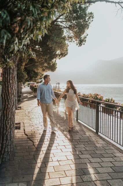 Lake Como engagement photoshoot along Menaggio’s tree-lined promenade The couple walk hand in hand along a tree-lined waterside path in Menaggio during their Lake Como engagement photoshoot.