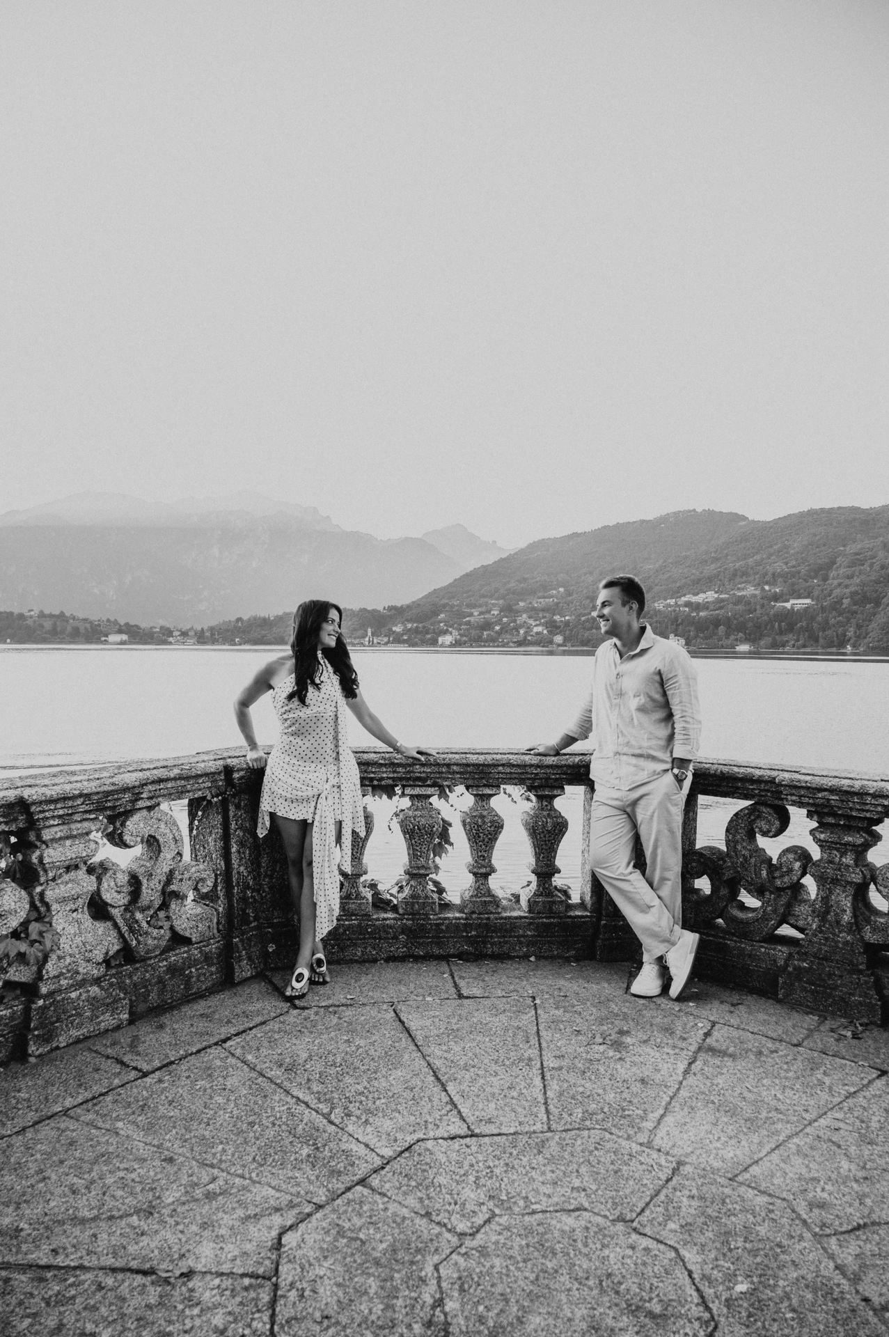 Black-and-white full-length photograph of the couple holding hands on a stone terrace during their Lake Como engagement photoshoot at Parco Civico Teresio Olivelli.