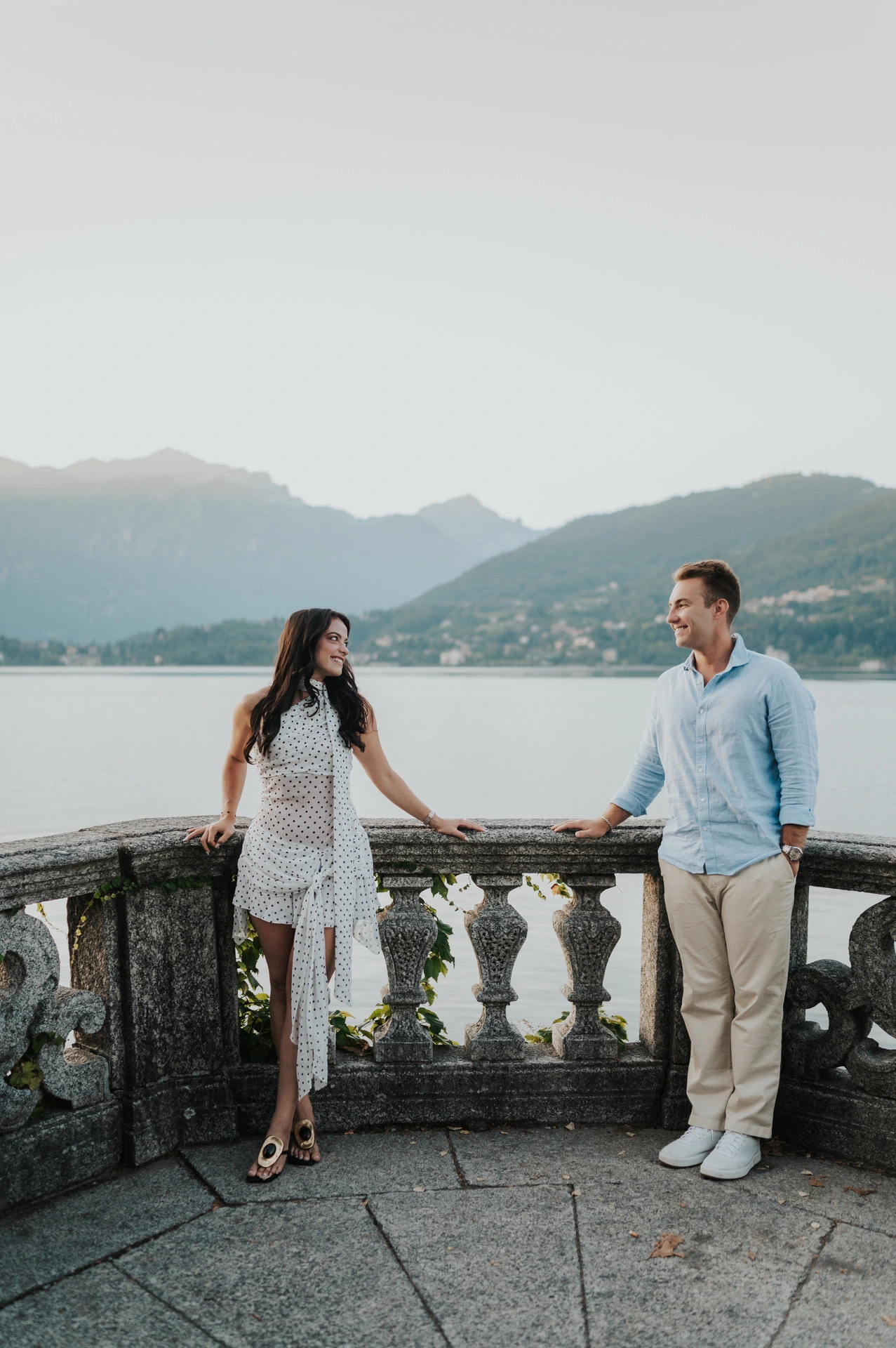 The couple hold hands across a stone balustrade corner, with the lake and mountains beyond during their Lake Como photoshoot at Parco Civico Teresio Olivelli.