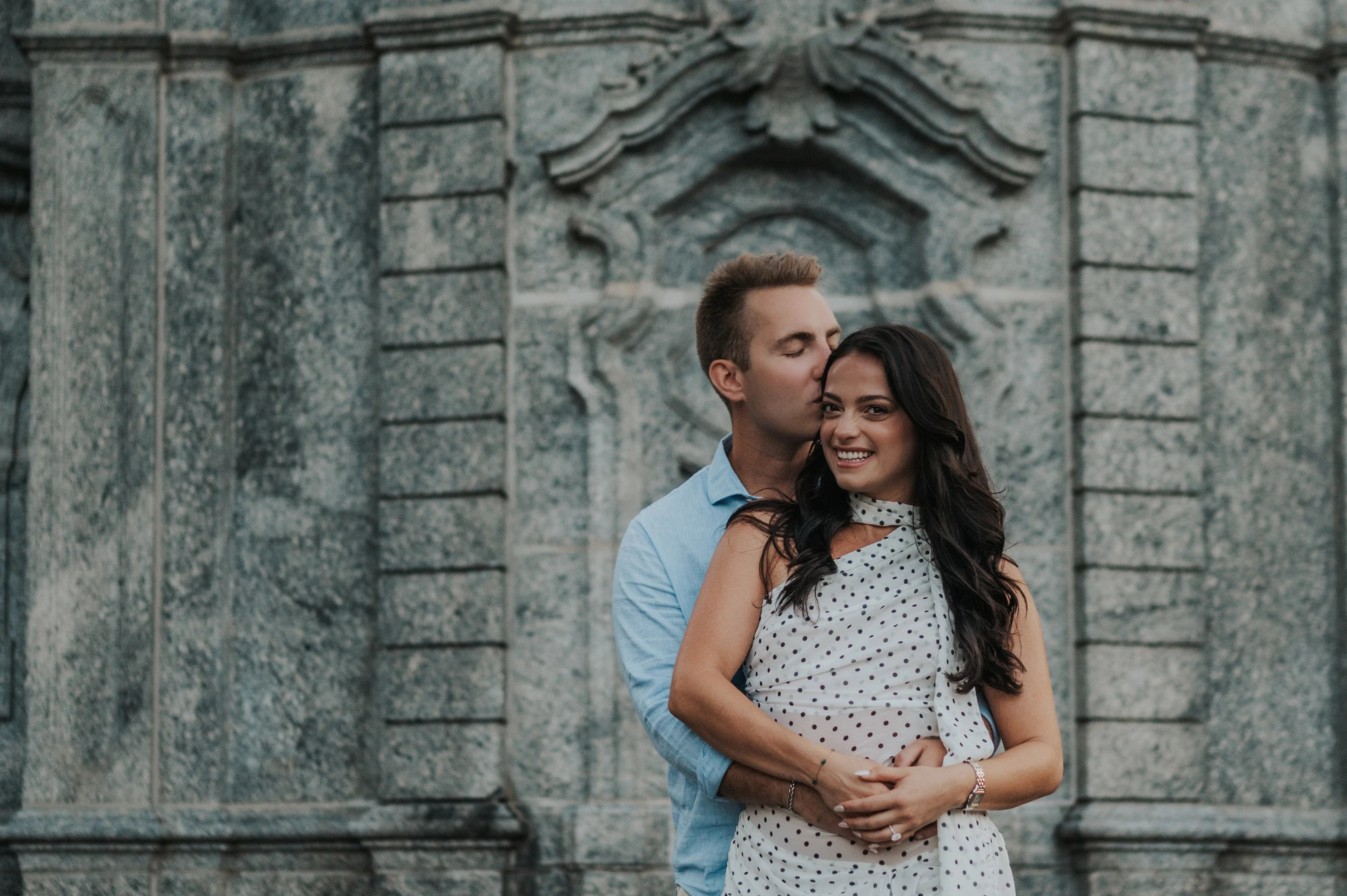 The man kisses the woman’s temple while they embrace before a carved stone wall during their Lake Como engagement photoshoot at Parco Civico Teresio Olivelli.