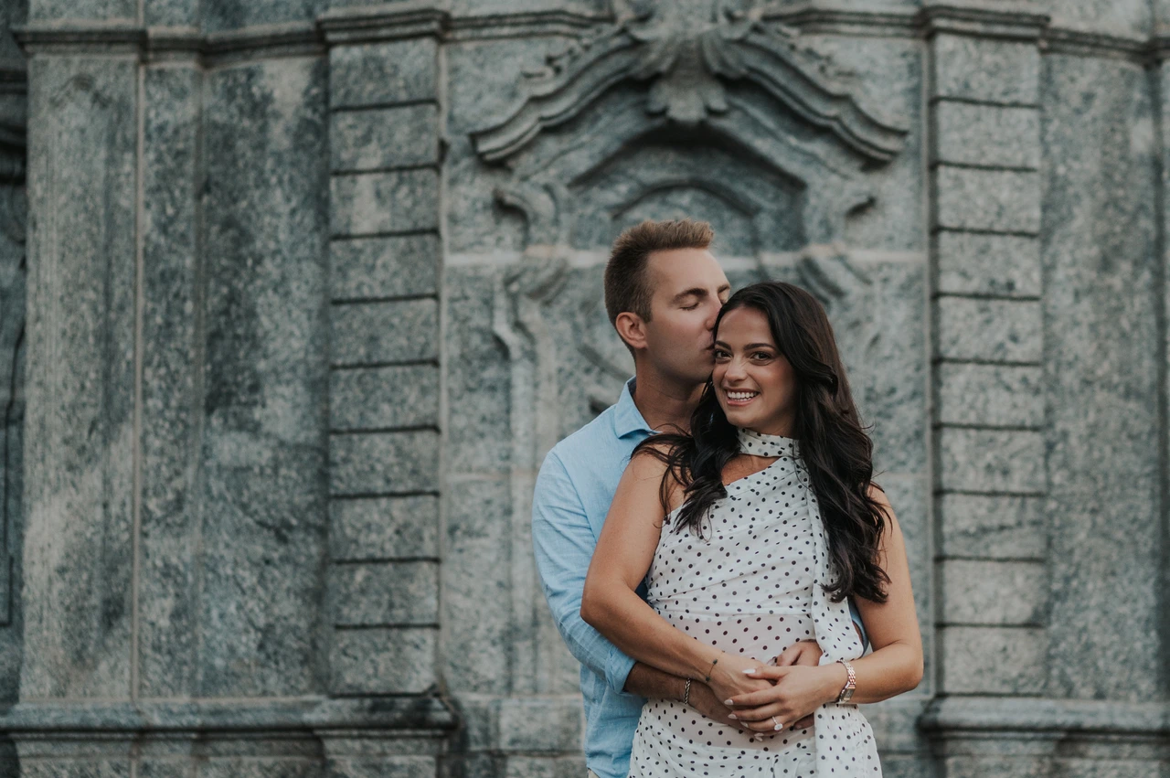 Lake Como engagement photoshoot against the park’s carved stonework The man kisses the woman’s temple while they embrace before a carved stone wall during their Lake Como engagement photoshoot at Parco Civico Teresio Olivelli.