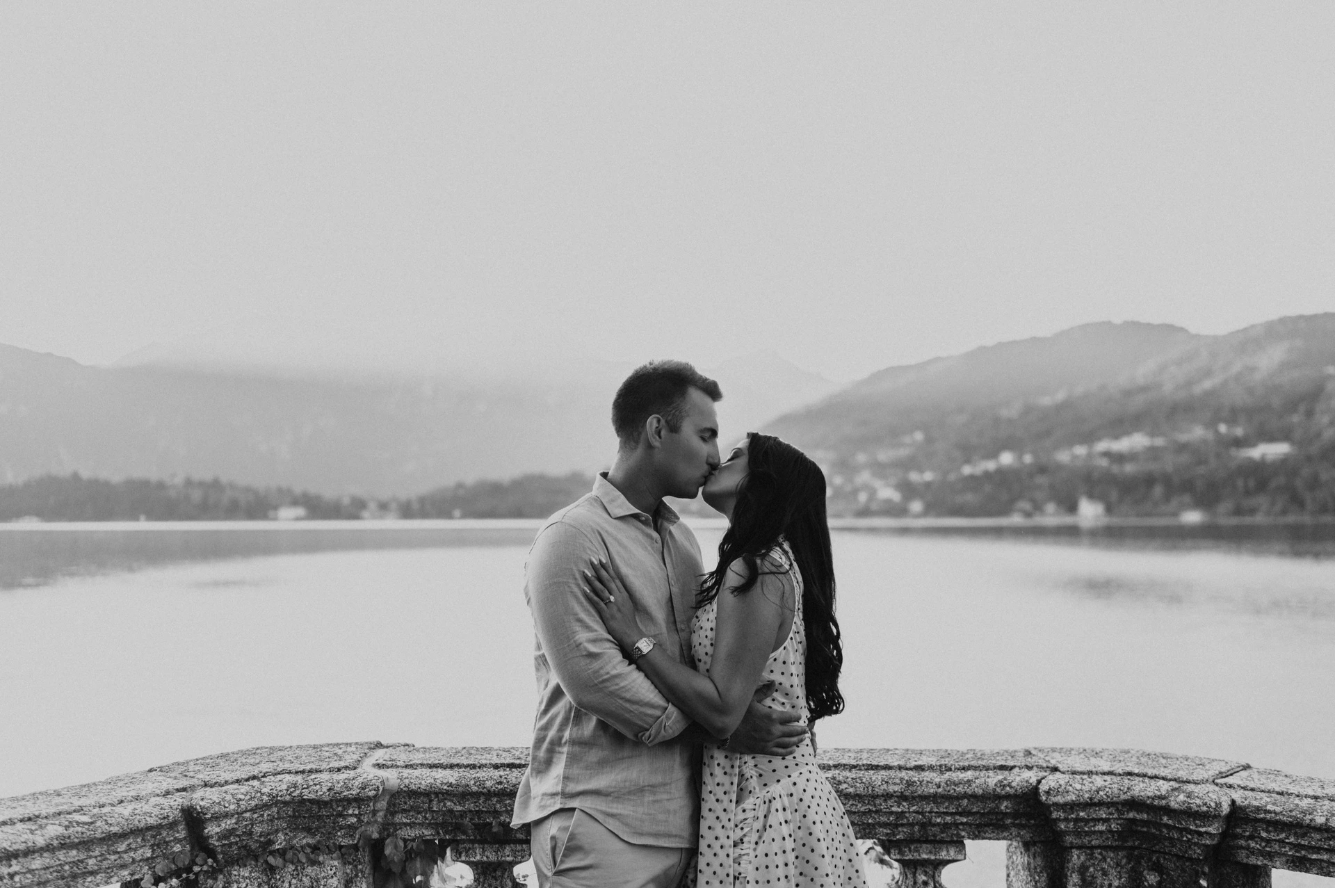 Black-and-white portrait of the couple holding each other beside the lake during their Lake Como engagement photoshoot at Parco Civico Teresio Olivelli.