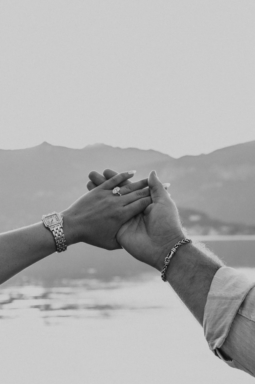 Black-and-white Lake Como engagement photoshoot with joined hands Black-and-white close-up of the couple’s joined hands raised against the lake and mountains during their Lake Como engagement photoshoot.