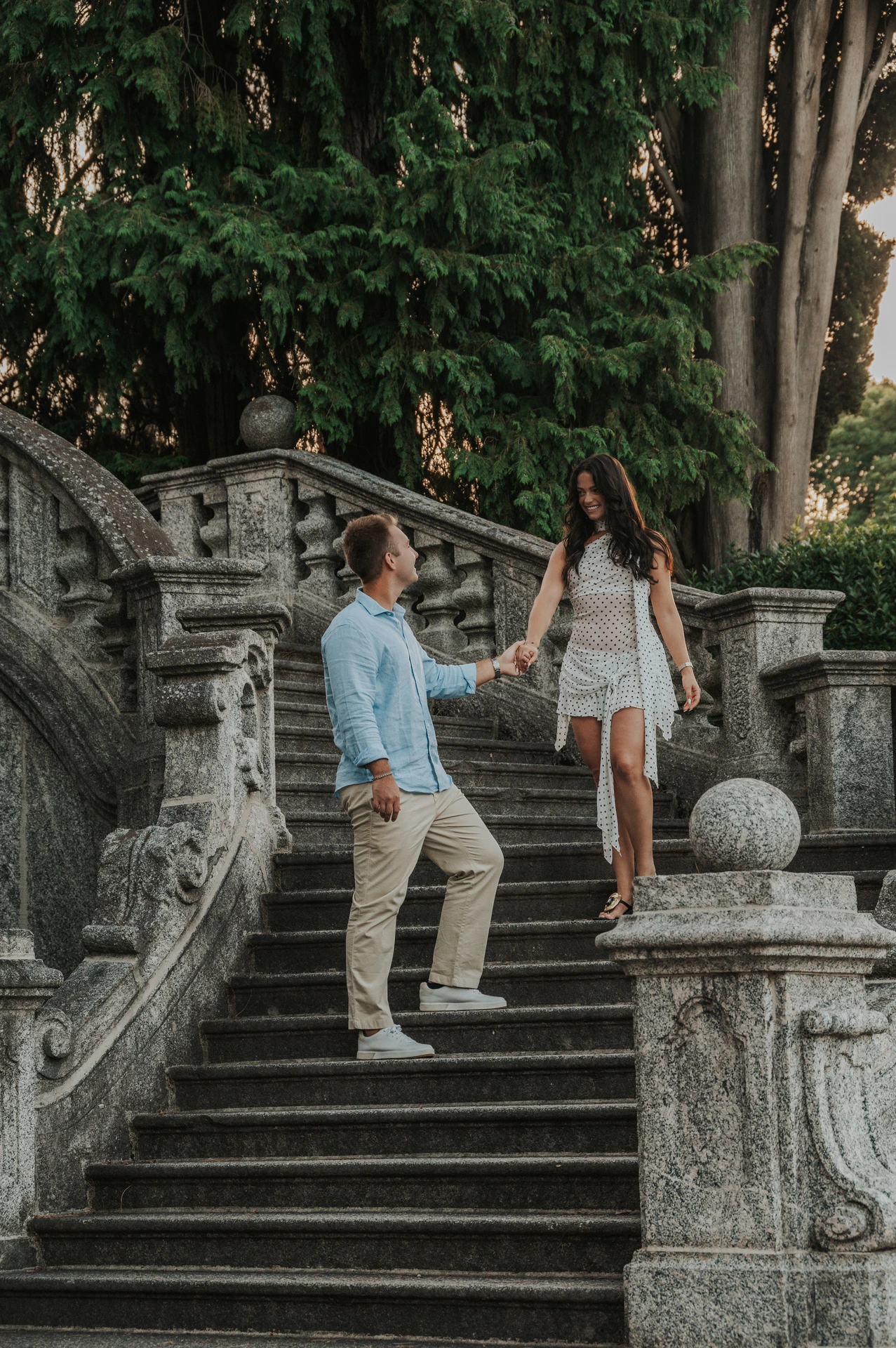 The couple hold hands across the stone steps beneath tall trees during their Lake Como engagement photoshoot at Parco Civico Teresio Olivelli.