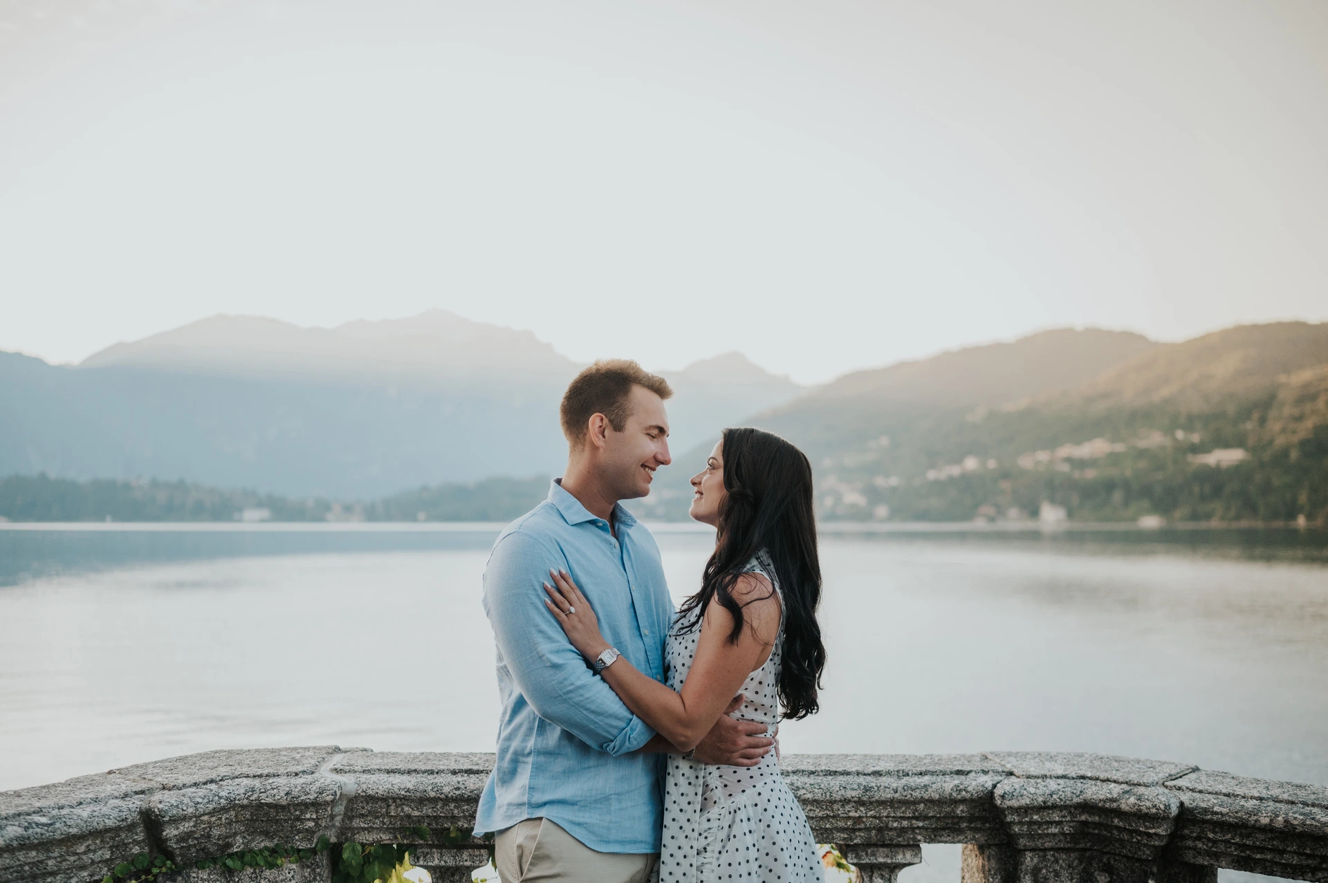 The couple hold each other and smile face to face beside the stone lake railing during their Lake Como engagement photoshoot.