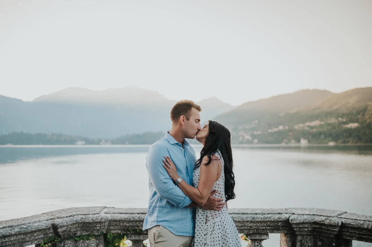 Lake Como photoshoot kiss beside the park’s lake terrace The couple kiss beside the stone balustrade with the water and distant hills behind them during their Lake Como photoshoot at Parco Civico Teresio Olivelli.