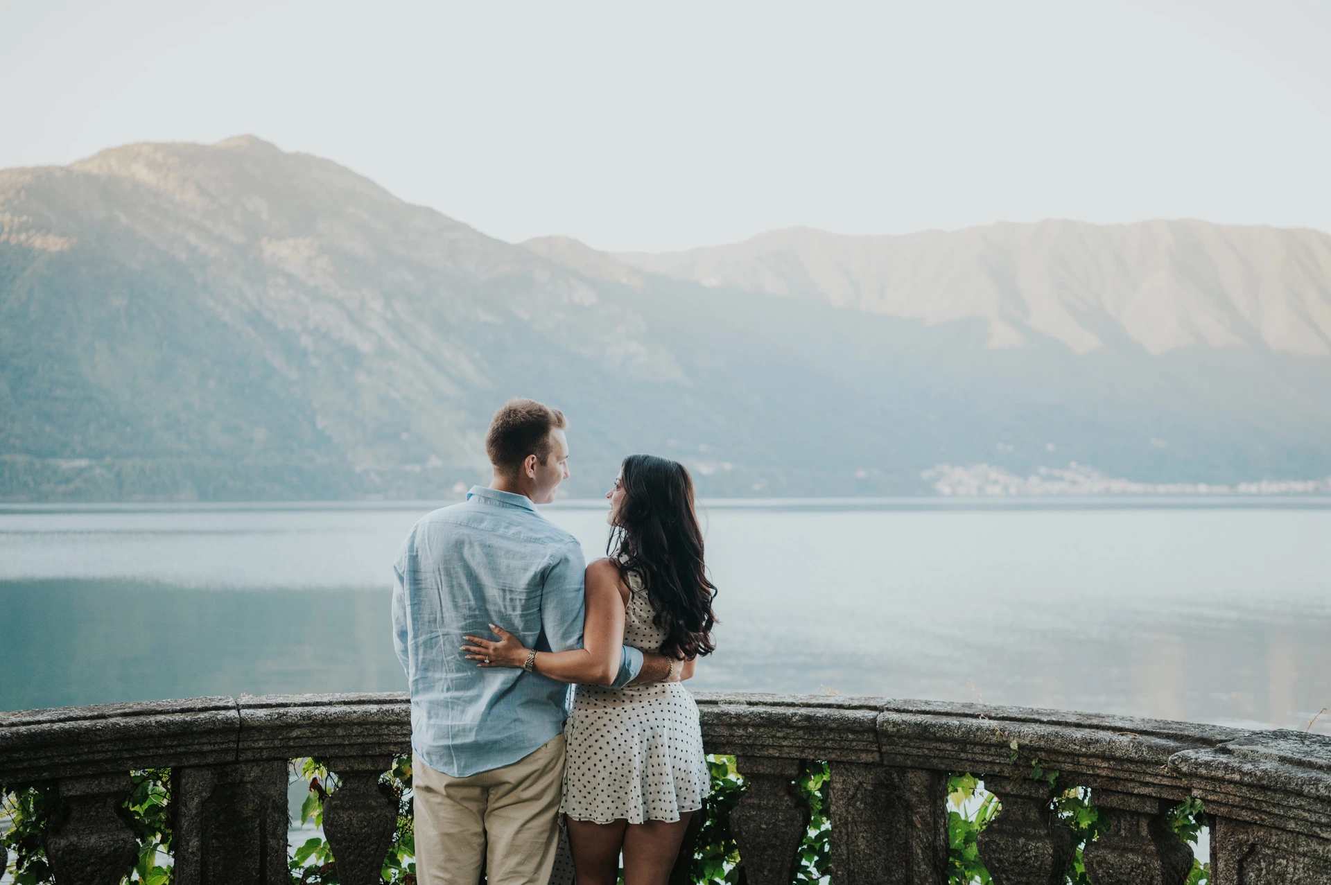 Seen from behind, the couple stand together at a stone balustrade looking over the water and mountains during their Lake Como photoshoot.