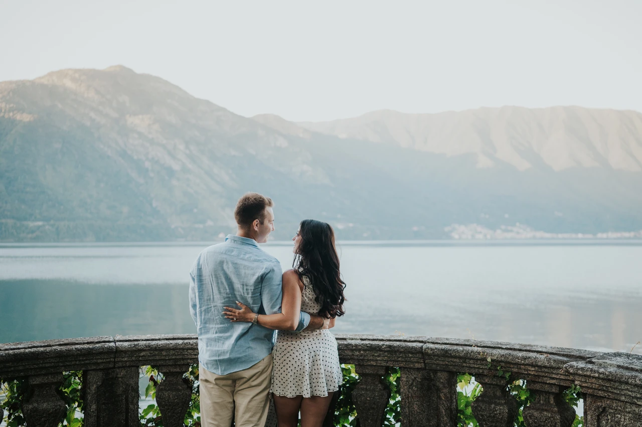 Lake Como photoshoot looking out from Parco Civico Teresio Olivelli Seen from behind, the couple stand together at a stone balustrade looking over the water and mountains during their Lake Como photoshoot.