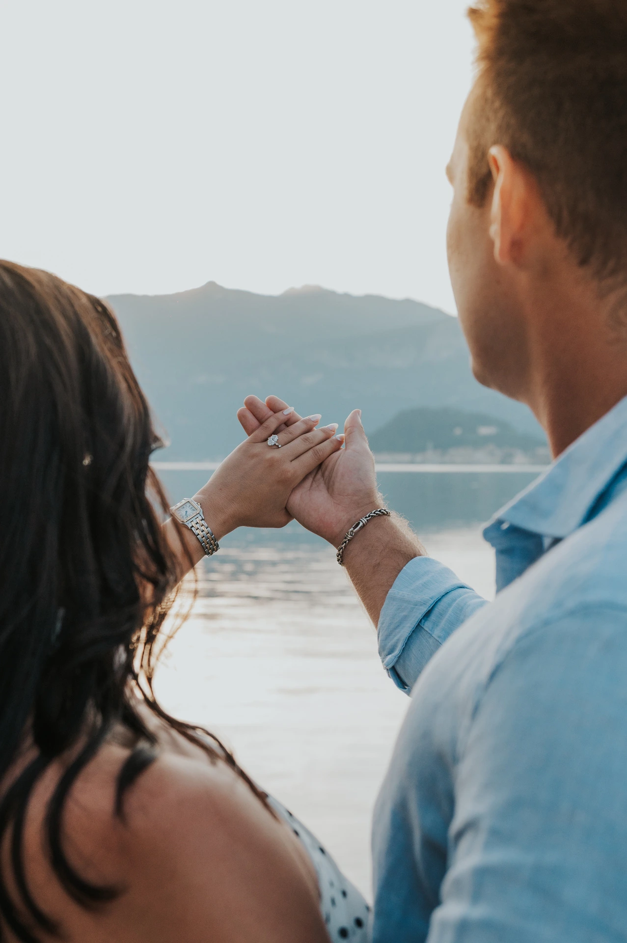 Seen over the couple’s shoulders, their joined hands and her ring are held against the lake view during their Lake Como photoshoot.