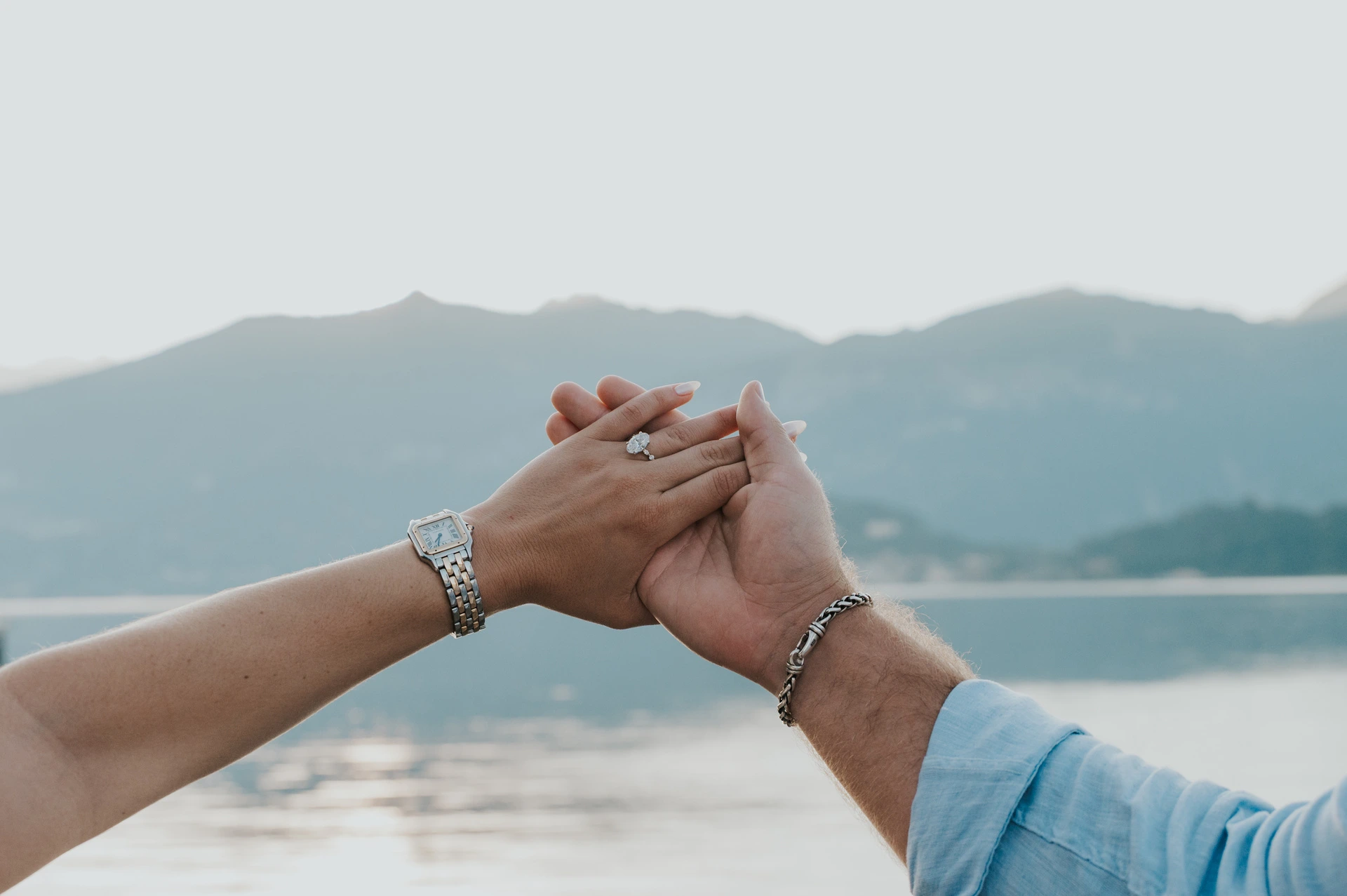A close view of the couple’s joined hands shows her engagement ring with the lake and mountains behind during their Lake Como photoshoot.