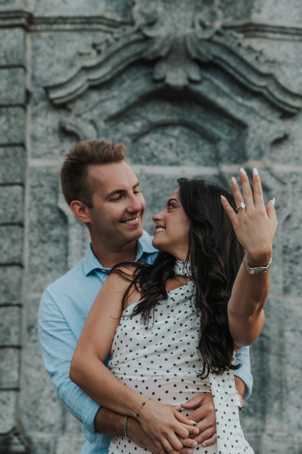 The woman holds her engagement ring towards the camera as the couple look at each other during their Lake Como photoshoot beside the park’s carved stone wall.