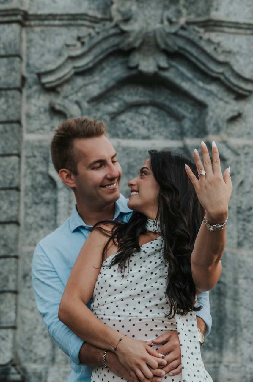 Lake Como photoshoot with the engagement ring before the stone wall The woman holds her engagement ring towards the camera as the couple look at each other during their Lake Como photoshoot beside the park’s carved stone wall.