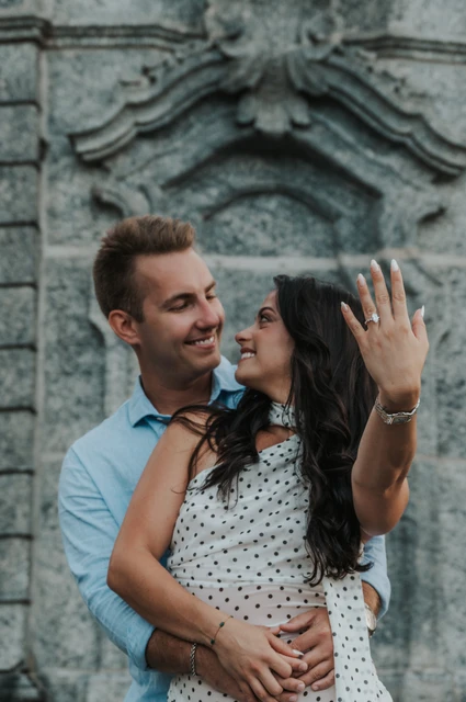 Lake Como photoshoot with the engagement ring before the stone wall The woman holds her engagement ring towards the camera as the couple look at each other during their Lake Como photoshoot beside the park’s carved stone wall.