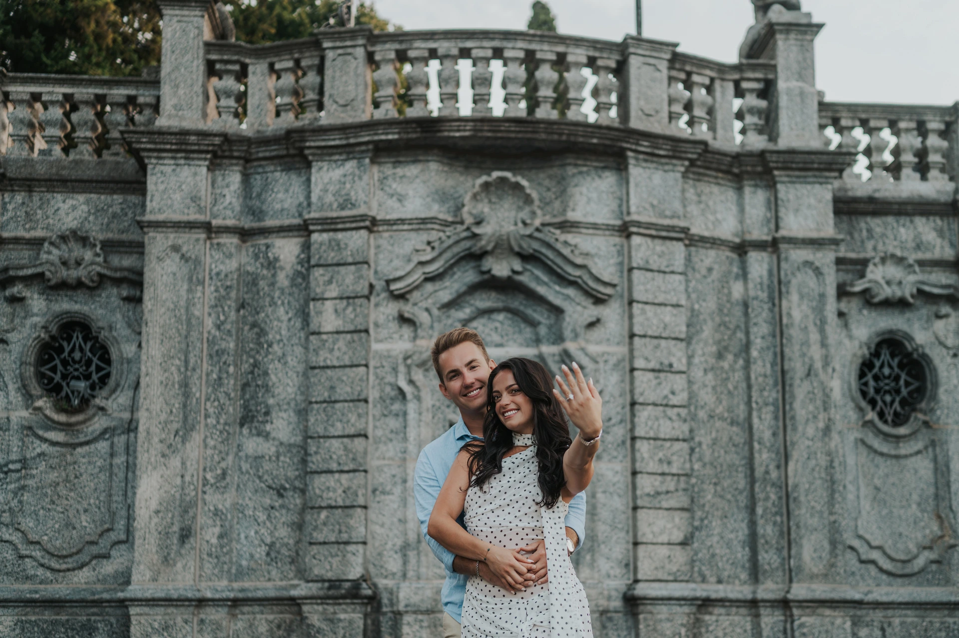 The woman shows her engagement ring while the couple stand together before an ornate stone wall during their Lake Como photoshoot.