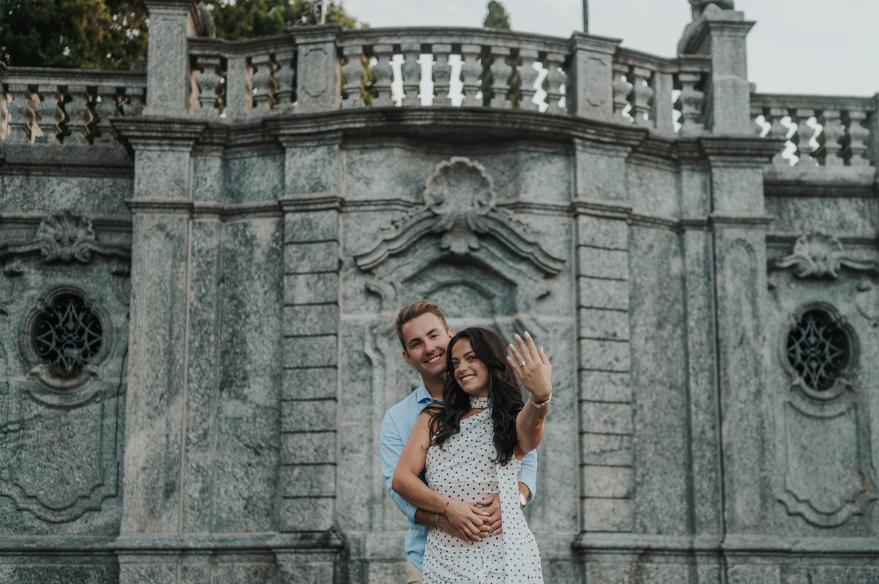 Lake Como engagement photoshoot showing the ring beside carved stone The woman shows her engagement ring while the couple stand together before an ornate stone wall during their Lake Como photoshoot.