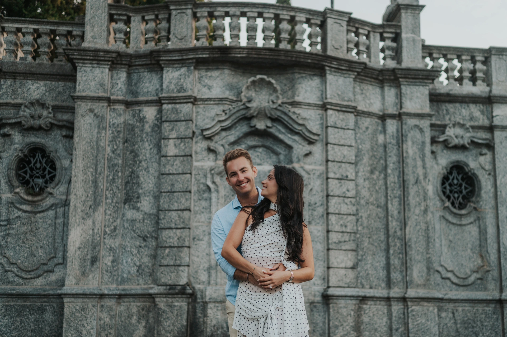 The couple smile together in front of decorative stone panels and a balustrade during their Lake Como photoshoot at Parco Civico Teresio Olivelli.